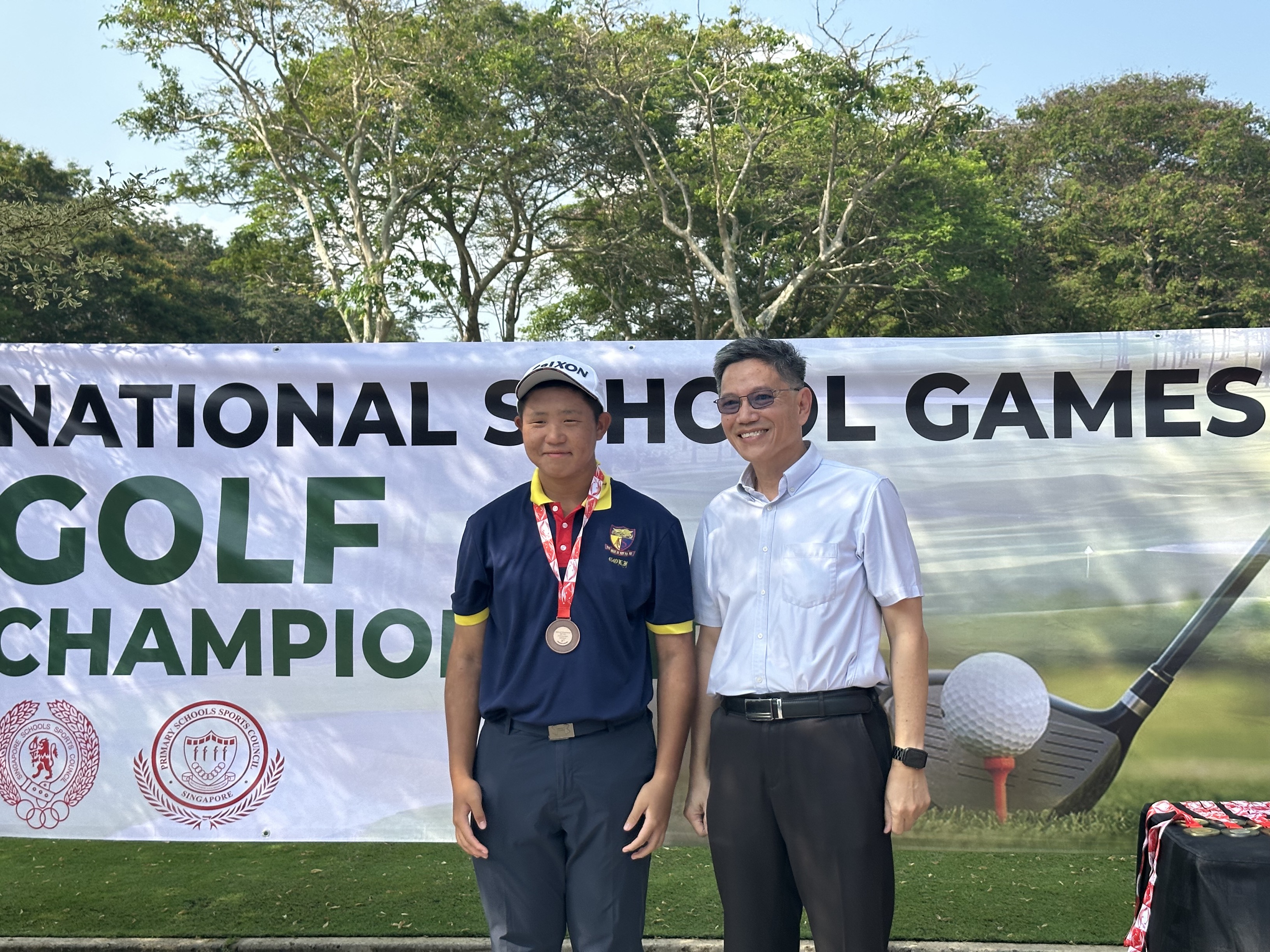 A young golfer wearing a medal poses with an older man in front of a National School Games Golf Championship banner.